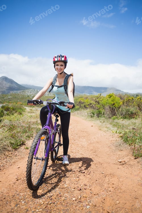 Preview: Woman standing on dirt trail holding purple mountain bike wearing cycling helmet and hydration pack
