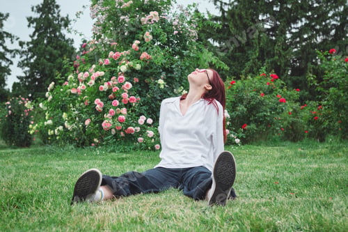 Preview: Teenage girl with red hair and glasses leaning back on grass in a blooming garden