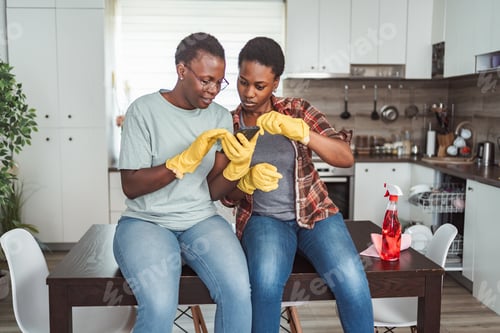 Preview: Two women wearing gloves using a smartphone while taking a break from cleaning the kitchen