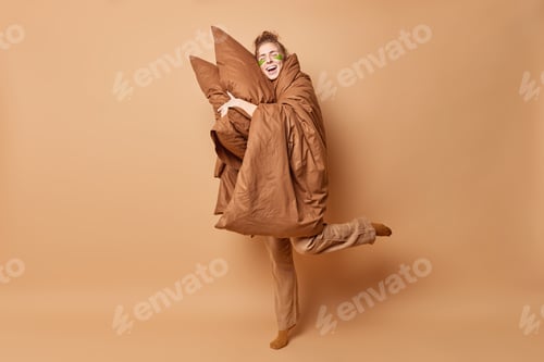 Preview: Studio shot of optimistic young woman leans on soft pillows takes nap wears pajama covered with blan