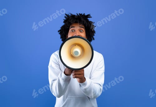 Preview: Emotional black teenager making announcement with megaphone on blue studio background