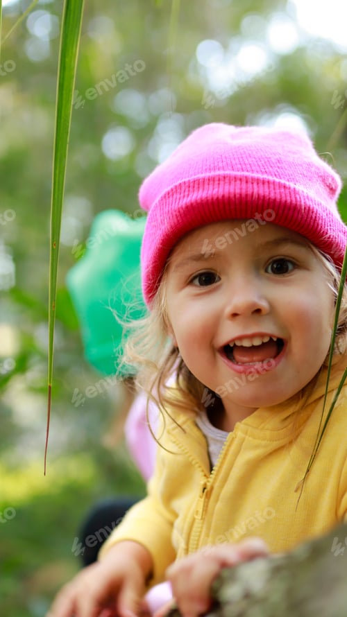 Preview: Happy little girl wearing yellow jacket and pink hat sitting on the tree branch