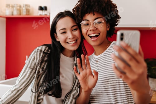Preview: Charming girls take selfie in kitchen. Woman in glasses holds smartphone and waves her hand