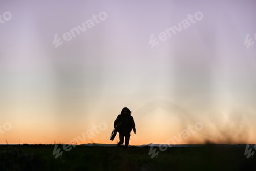 Preview: silhouette of a caucasian boy with long hair holding a guitar running towards camera at sunset