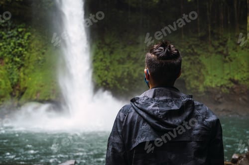 Preview: Back view of a male traveler wearing a blue mask enjoying the view of La Fortuna waterfall