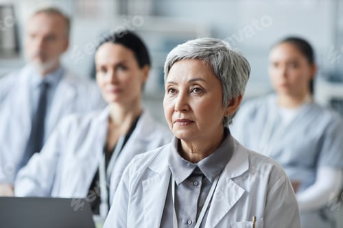 Preview: Mature Asian female doctor in lab coat looking at speaker making report