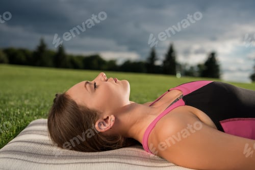 Preview: side view of young woman resting on yoga mat after practicing yoga in park