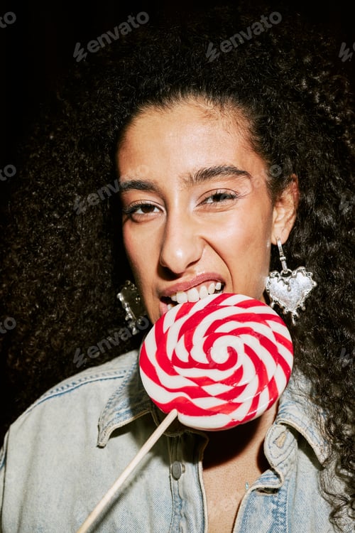 Preview: Portrait of Young Adult Middle Eastern Woman Biting Swirled Lollipop in Booth