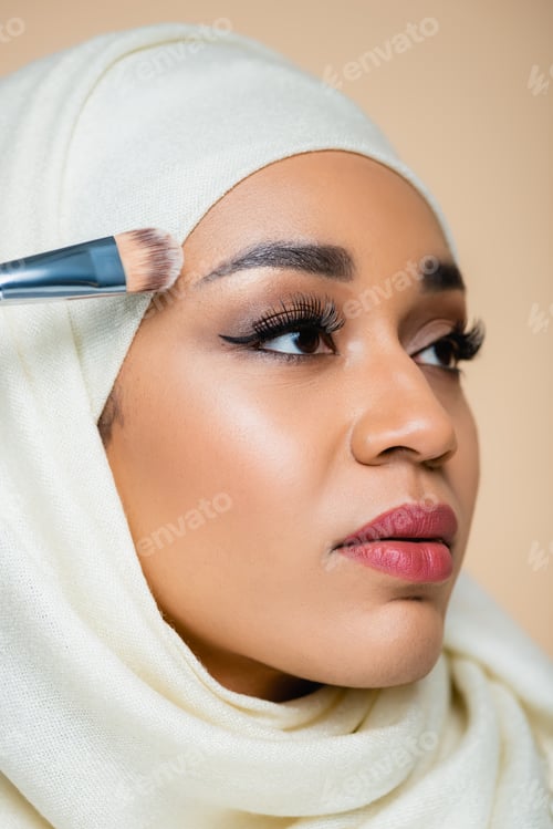 Preview: portrait of young muslim woman in hijab applying face foundation with cosmetic brush isolated on