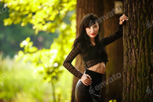 Preview: sexy brunette girl in a lace blouse and leather skirt with high heels posing near tree in the woods