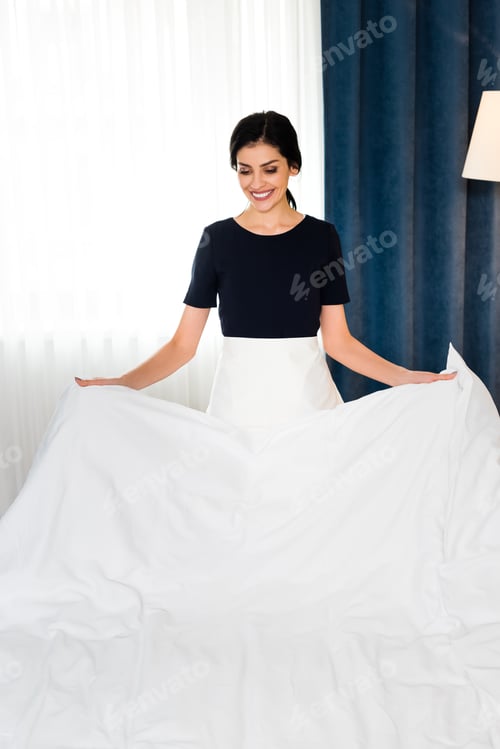 Preview: happy housemaid holding white bed sheet in hotel room
