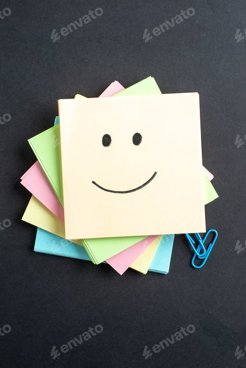 Preview: Vertical view of smiling emoji on one of stacked sticky note cubes paper clips on isolated black