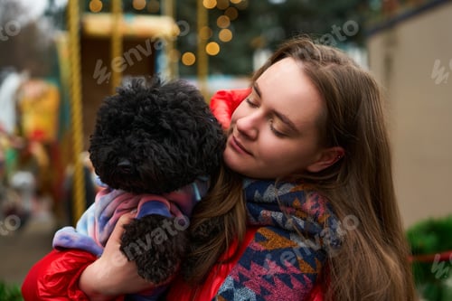 Preview: Young woman enjoys winter outing, holding fluffy black dog in warm scarf amidst festive decorations