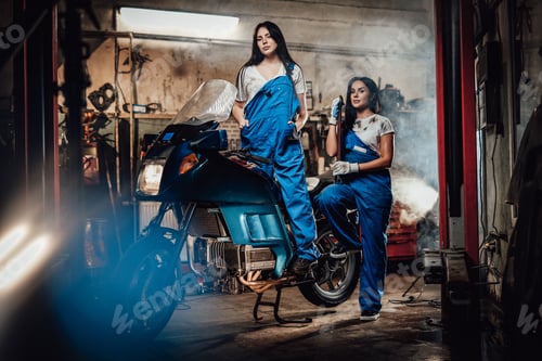 Preview: Two brunette women in blue overalls posing next to a sportbike in authentic workshop garage