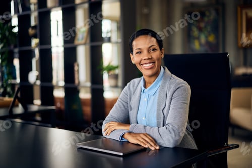 Preview: Portrait of a smiling black woman, posing for the camera while sitting at the office.