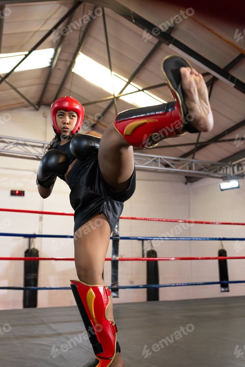 Preview: Female boxer kicking right leg, wearing red headgear, black gloves and shin guards in boxing ring