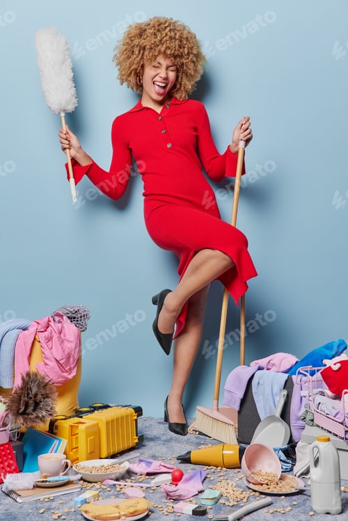 Preview: Vertical shot of cheerful curly haired woman holds broom and dusting brush wears long red dress does