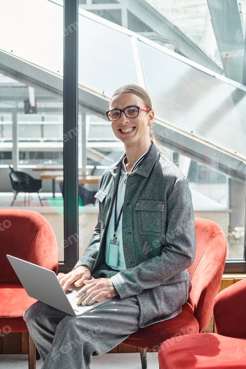 Preview: smiling young employee with glasses looking at camera while working on his laptop, coworking