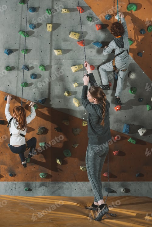 Preview: Full-length shot of two little kids climbing a wall at gym and their mother holding securing rope