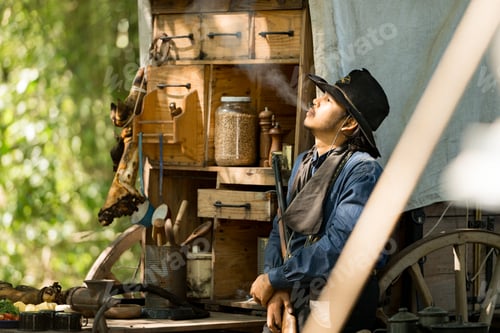 Preview: A cowboy stands holding a rifle in a forest camp, with sunlight streaming through the smoke