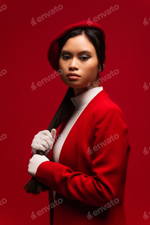Preview: Brunette asian model in beret touching hair isolated on red