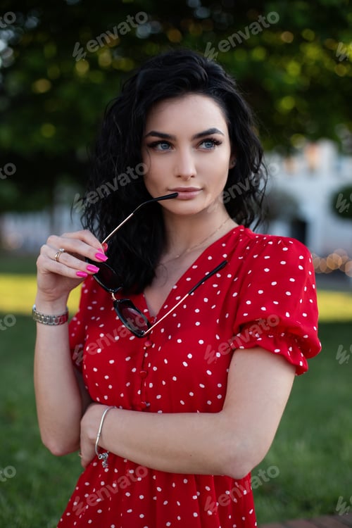 Preview: Elegant beautiful young curly woman in red vintage dress holding sunglasses and walks in the town