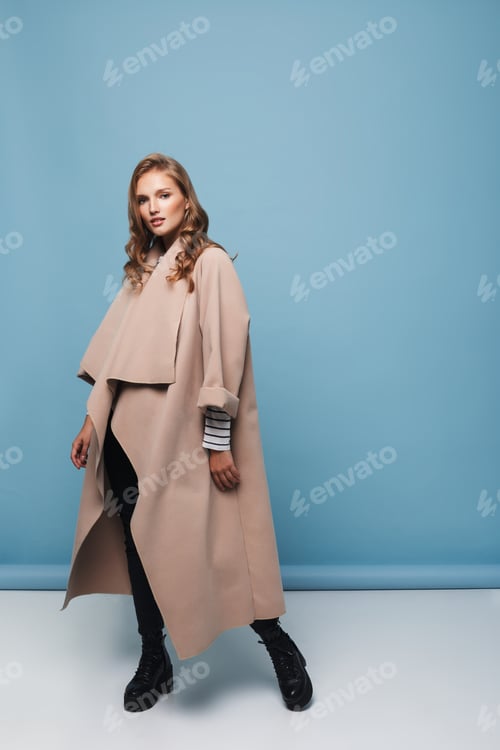 Preview: Young beautiful woman with wavy hair in beige coat and black boots dreamily looking in camera