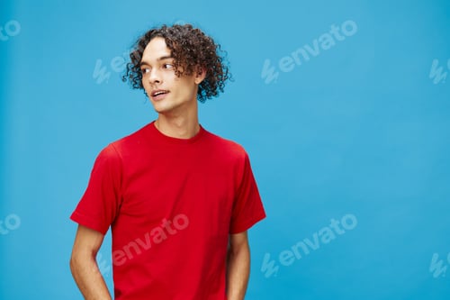 Preview: Attractive cute young curly tanned Caucasian guy in basic red t-shirt looks aside posing isolated on