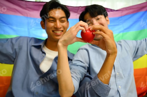 Preview: Loving gay couple making a heart shape with their hands and pride rainbow flag behind them