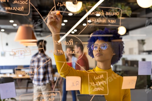 Preview: Biracial casual businesswoman with blue afro making notes on glass wall in office