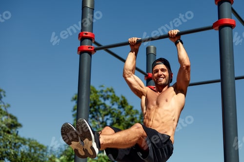 Preview: Athletic young man hanging from the bars at the calisthenics gym outdoors smiling
