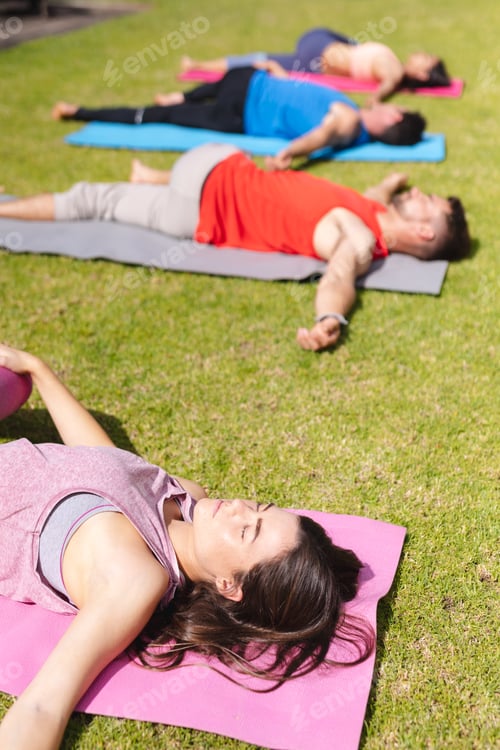 Preview: Young woman lying on exercise mat with eyes closed while practicing yoga with people in park