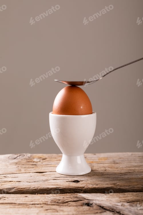 Preview: Close-up of spoon on fresh boiled brown egg in cup on table against gray background with copy space