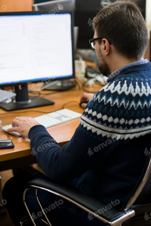 Preview: Young man with glasses sits in the office in front of his laptop and monitor working on it