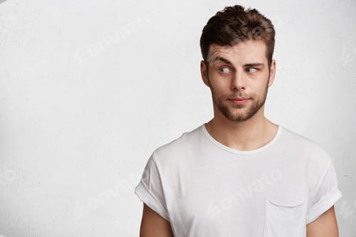 Preview: Young bearded attractive male with skeptic look, raises eyebrow in bewilderment, has some doubts and