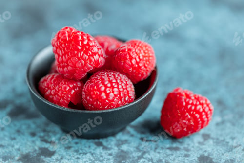 Preview: Fresh ripe raspberries in a small black bowl on a textured blue table. Macro photography.