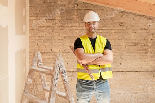 Preview: Male Construction worker in safety hardhat posing with level tool and crossed arms at building site