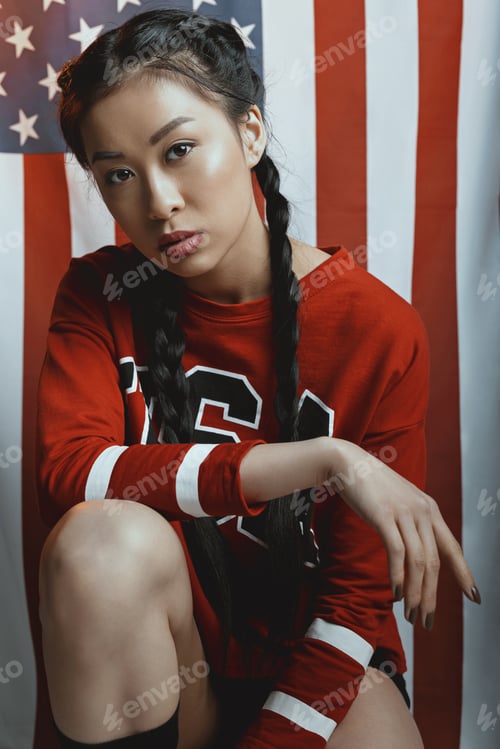 Preview: Hipster asian girl in american patriotic outfit with braids looking at camera with us flag on