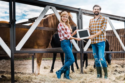 Preview: smiling couple of farmers holding blackboard in stable