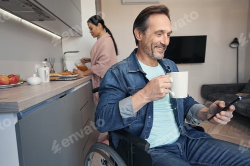 Preview: Smiling male using wheelchair and smartphone in kitchen with wife
