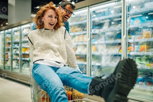 Preview: Young couple having fun while shopping in supermarket. Woman sitting in shopping cart