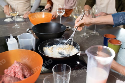 Preview: Hands of mature woman mixing up milk and butter in frying pan against other trainees cooking