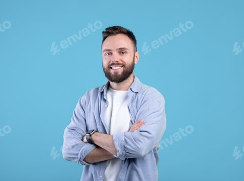 Portrait of positive young guy posing with crossed arms and smiling at camera on blue studio