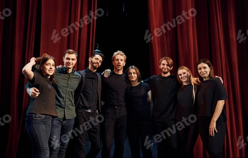 Preview: Standing against red curtains. Group of actors in dark colored clothes on rehearsal in the theater