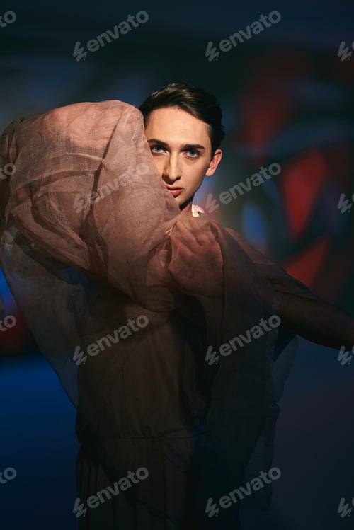 Preview: well-dressed androgynous fashionista in pastel attire looking at camera on vibrant backdrop