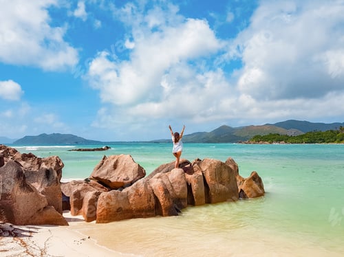 Preview: Rear view of woman standing on rock formation on tropical sandy beach.