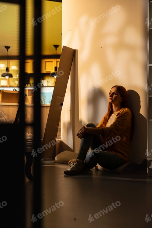 Preview: Tired and worried woman sitting indoors surrounded by moving boxes
