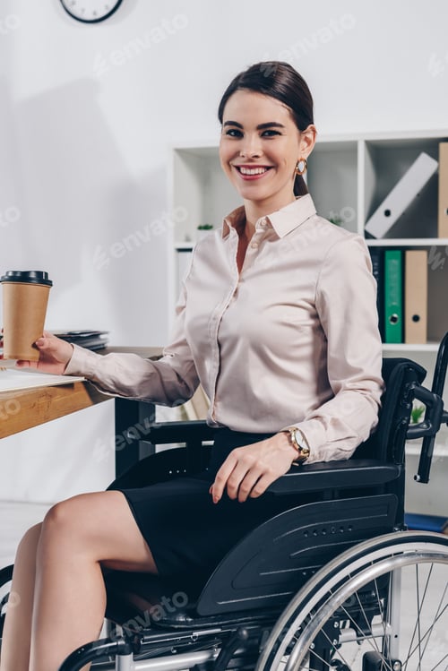 Preview: Disabled employee on wheelchair looking at camera and smiling with paper cup of coffee in office
