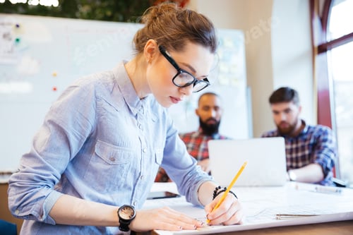 Preview: Woman Engineer Working On Blueprint In Office With Colleagues On Background