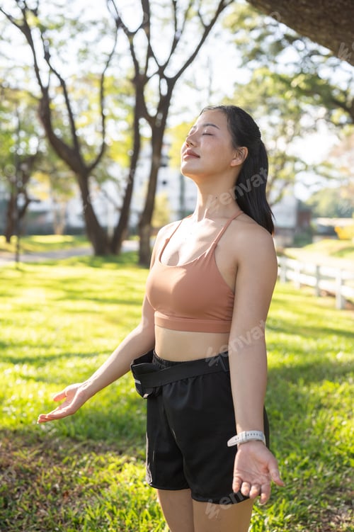 Preview: Pretty asian woman in sportswear closed eyes open arms standing or resting after exercise in park.